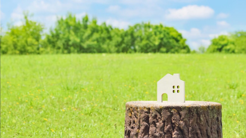 一個小型白色房子模型放置在樹樁上，背景是綠色的草地和藍天，陽光明媚。